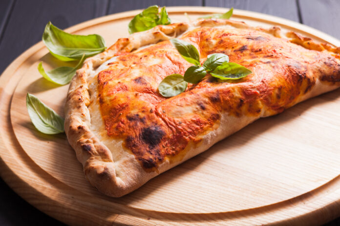 Calzone caseiro dourado servido em uma tábua de madeira com folhas de manjericão fresco.