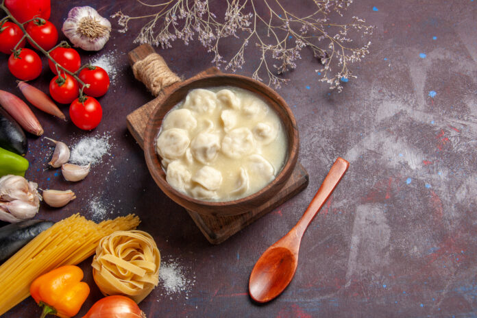 Tigela de madeira com molho bechamel cremoso acompanhando tortellini, cercada por ingredientes frescos como tomates, alho, pimentão e massa em uma superfície rústica.