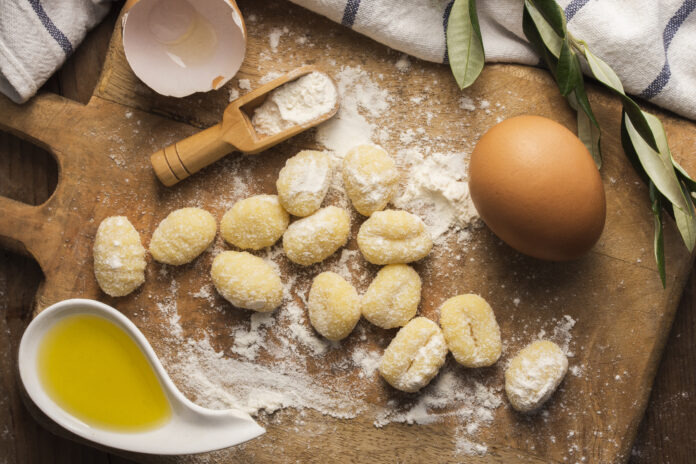 Ingredientes para preparar gnocchi caseiro, incluindo ovos, farinha e azeite, em uma tábua de madeira.