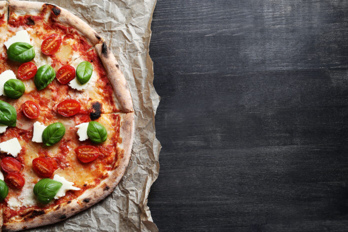 Pizza italiana caseira com folhas de manjericão fresco, queijo e tomates-cereja, sobre papel de forno em uma superfície rústica escura.