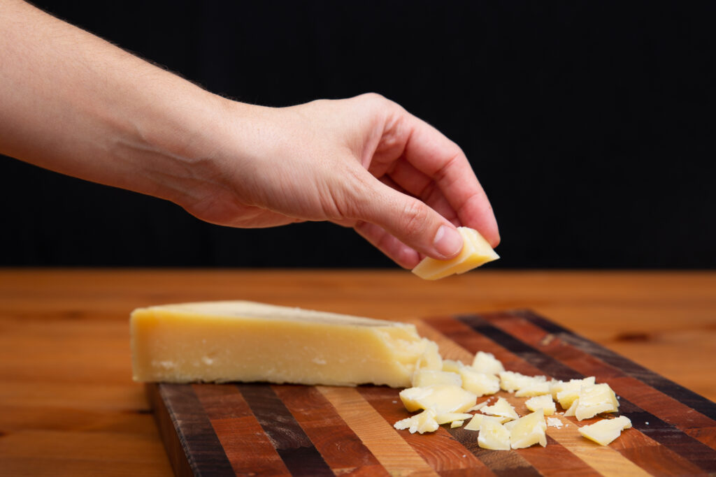 Mão segurando um pedaço de queijo Pecorino sobre uma tábua de madeira com lascas de queijo.