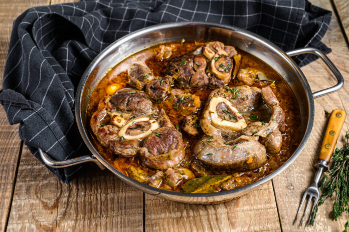 Panela de ossobuco alla milanese com caldo aromático e ervas frescas sobre uma mesa rústica de madeira.