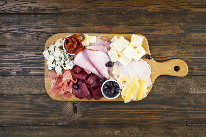 Tábua de frios italiana com variedade de queijos, presunto, salame, azeitonas e pimentões secos, disposta em tábua de madeira rústica.