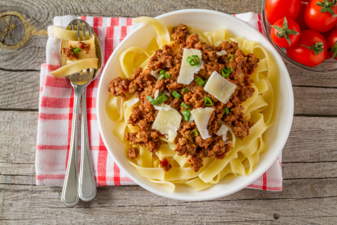 Prato de tagliatelle ao ragù servido em um bowl branco, com molho de carne, lascas de queijo parmesão e cebolinha, acompanhado de talheres sobre uma toalha xadrez vermelha e branca.