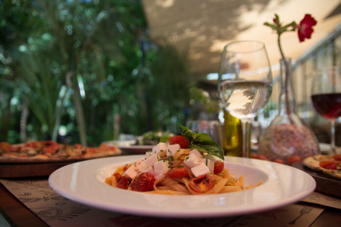 Prato de massa italiana com tomates frescos, manjericão e queijo, acompanhado por taças de vinho branco e uma atmosfera acolhedora ao ar livre.