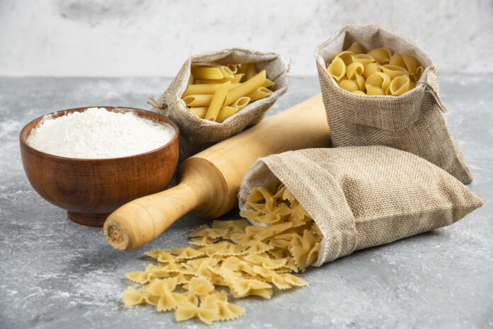 Sacos de massas secas e farinha sem glúten com um rolo de madeira sobre uma superfície cinza.