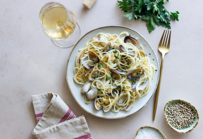 Prato de spaghetti alle vongole servido com vôngoles frescos, alho, salsinha e acompanhado de uma taça de vinho branco, ideal para harmonização.