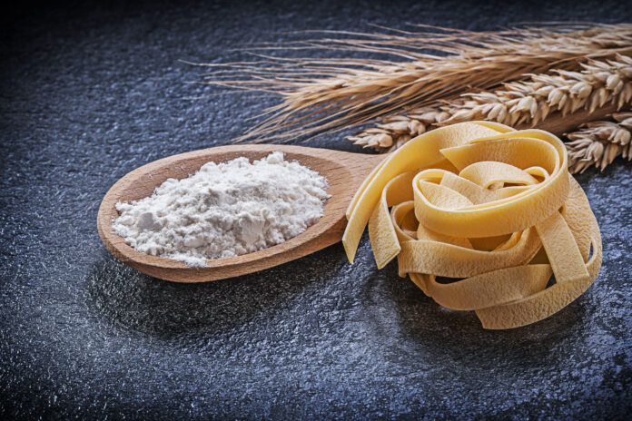 Farinha de trigo italiana em uma colher de madeira, com massa fresca e espigas de trigo ao fundo.