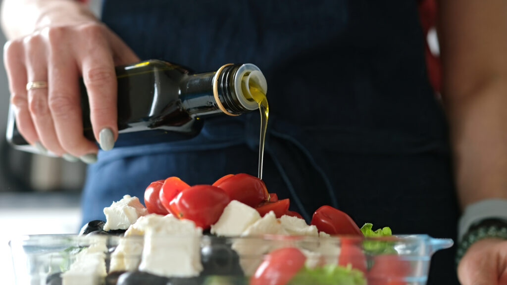 Pessoa despejando azeite italiano extra virgem sobre uma salada de tomates, queijo feta e azeitonas.