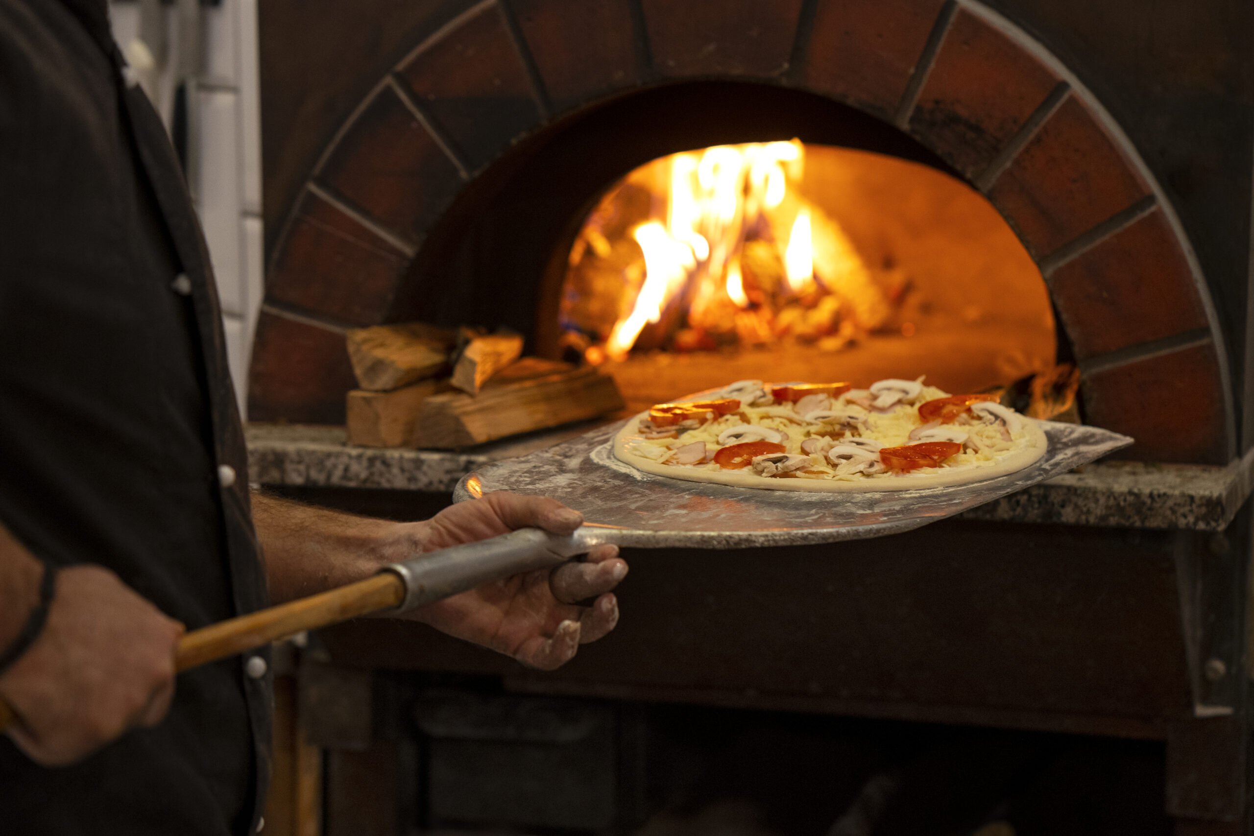 Pizza sendo inserida em um forno a lenha com ingredientes frescos e uma chama viva ao fundo.