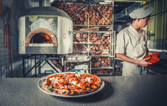 Pizza italiana fresca com cobertura tradicional em uma pizzaria com forno a lenha ao fundo.